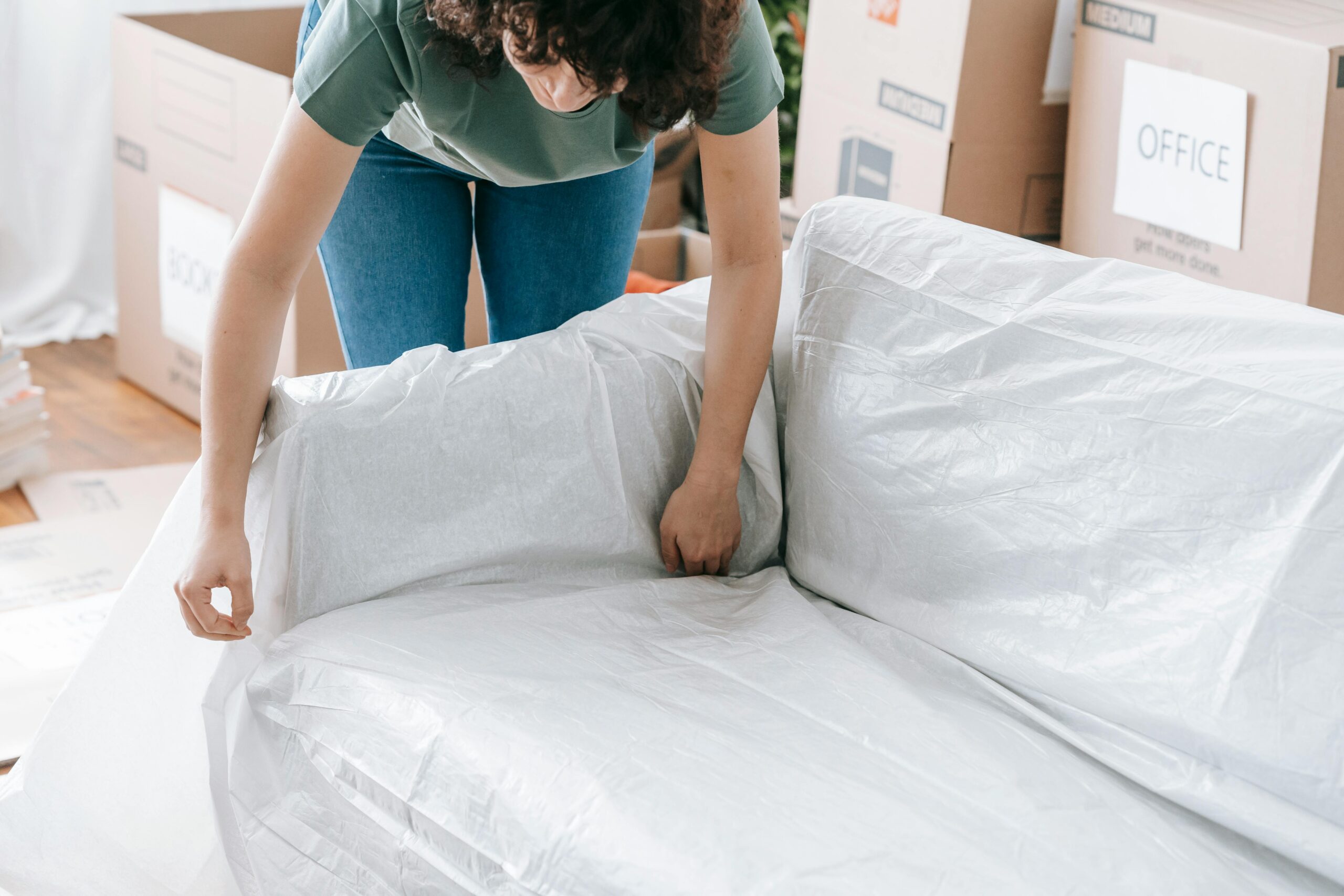 A woman carefully wraps a sofa in protective material while packing boxes in a bright room.